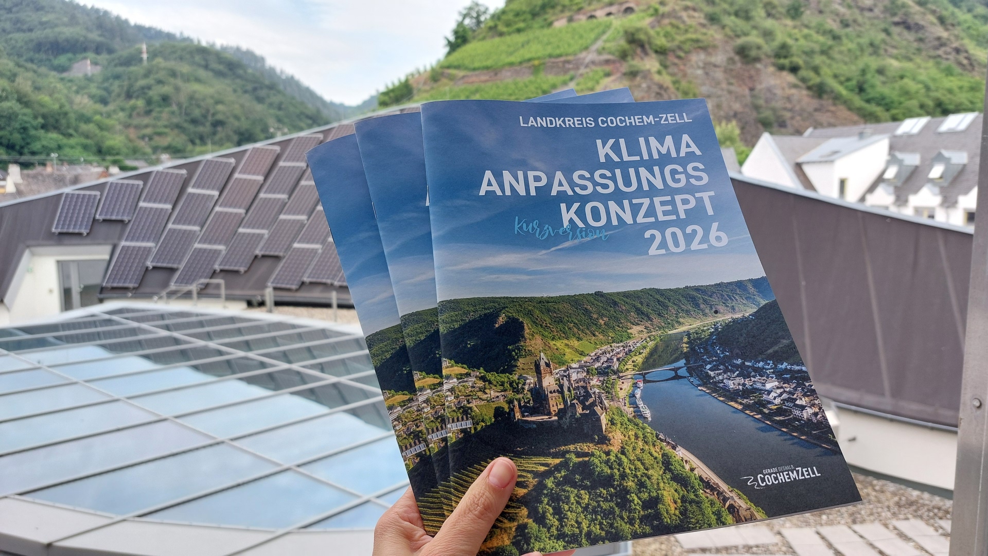 Hand hält drei Broschüren "Klimaanpassungskonzept Kurzversion" hoch. Im Hintergrund eine Dachfläche mit PV-Anlage und grüne Berge.