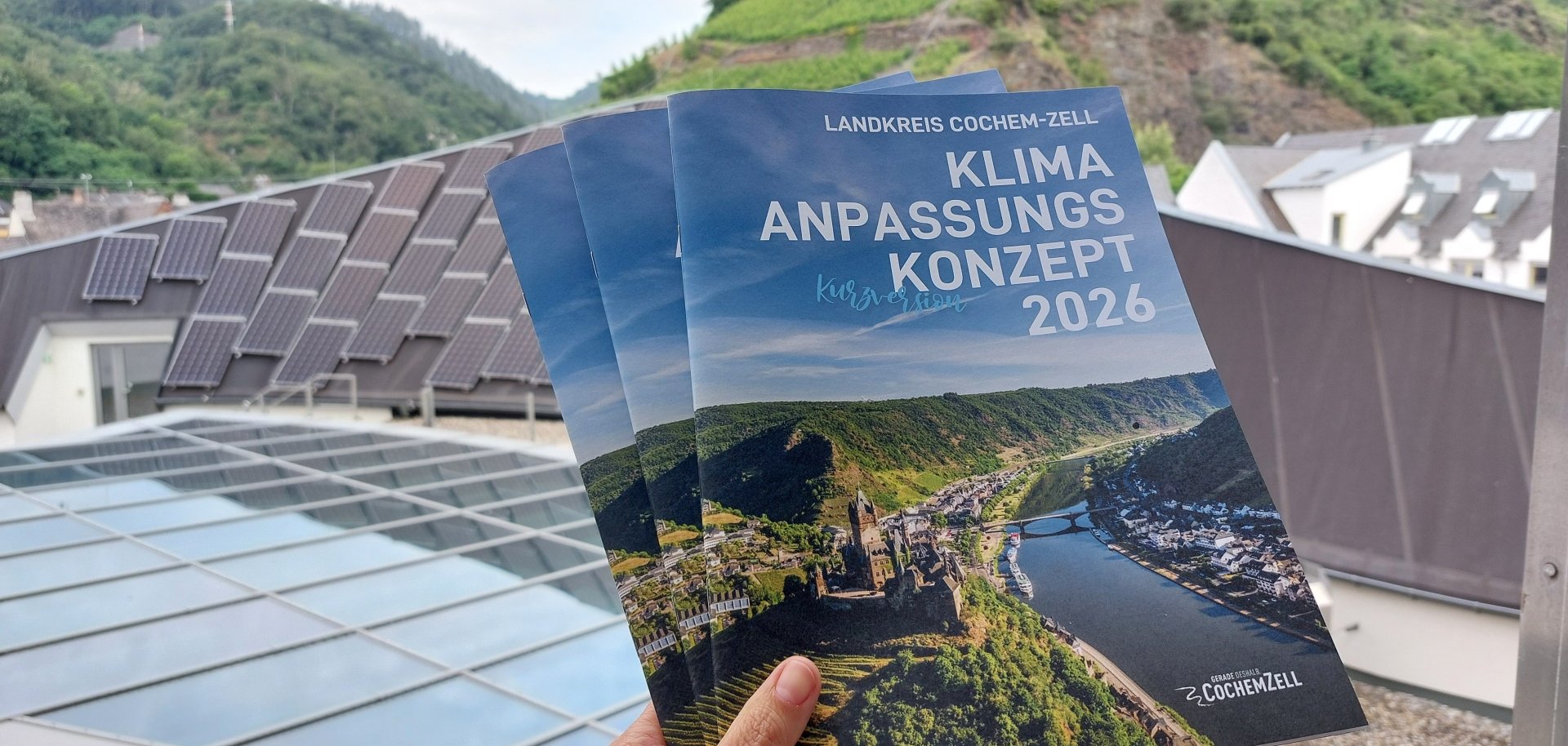 Hand hält drei Broschüren "Klimaanpassungskonzept Kurzversion" hoch. Im Hintergrund eine Dachfläche mit PV-Anlage und grüne Berge.